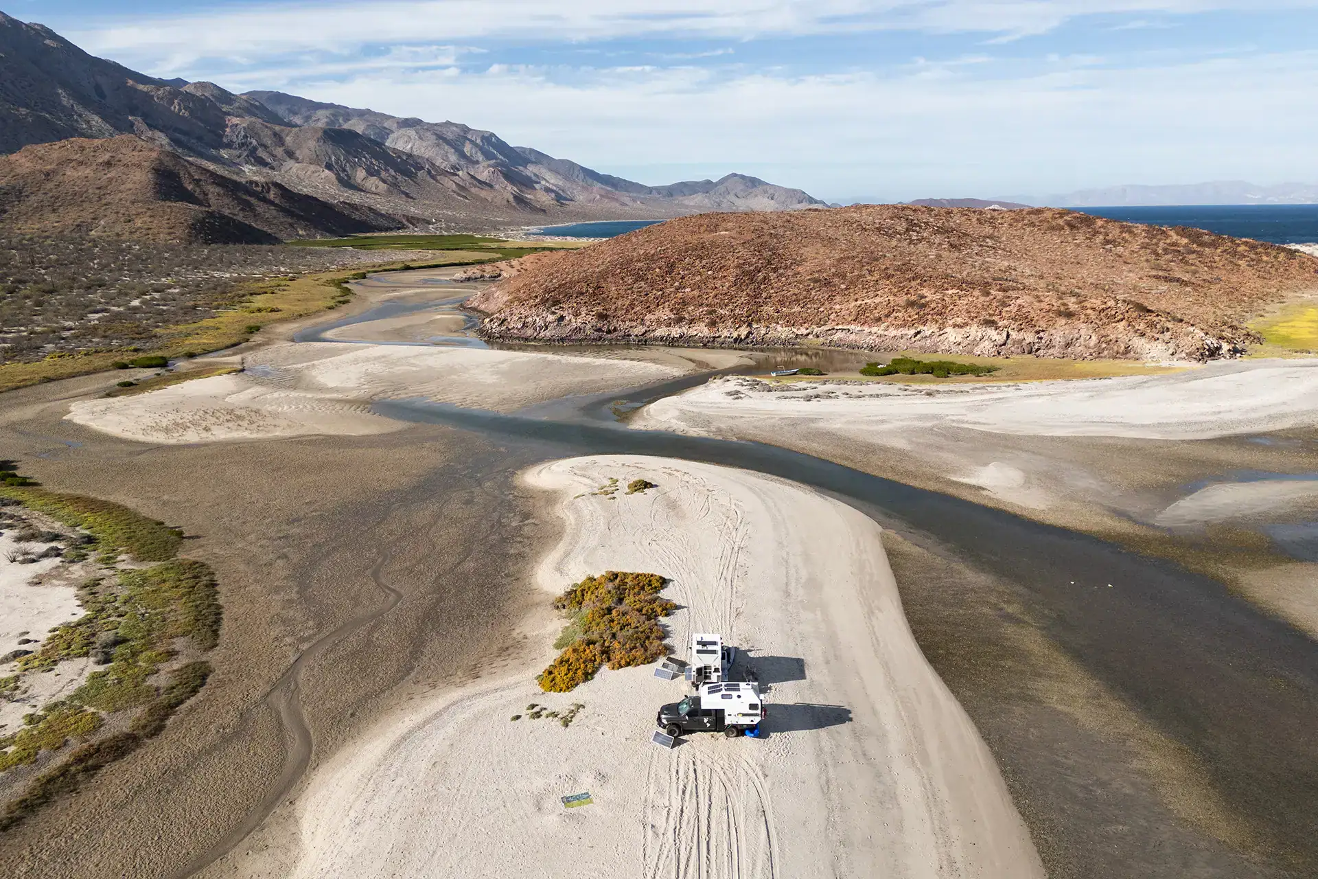 Four Wheel Camper vans boondocking off-grid with solar panels, overlanding remote sandy landscape, mountains, river, ocean.