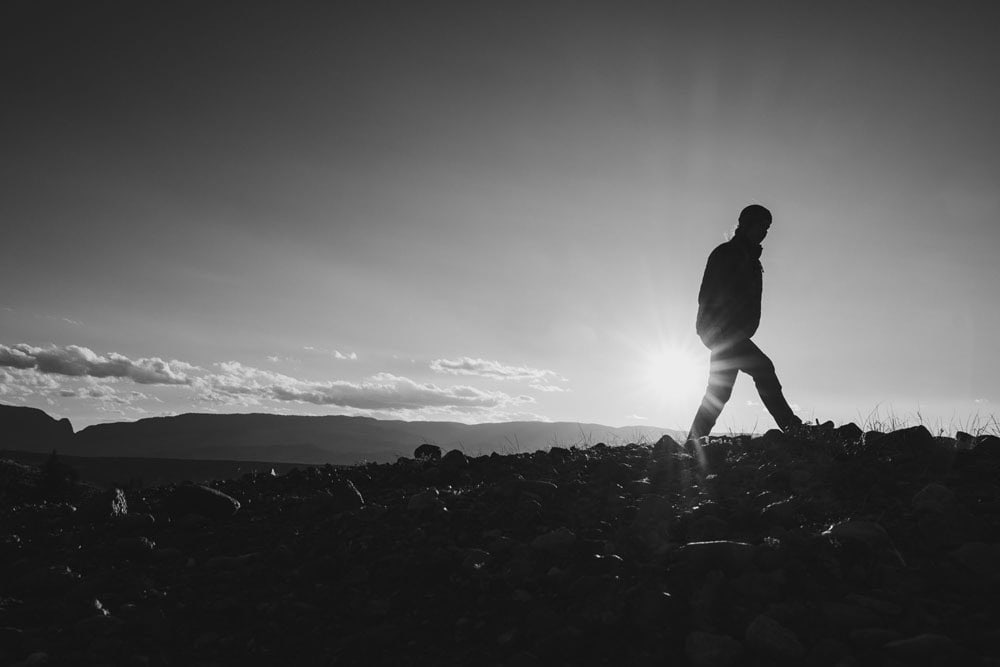 Silhouette of person walking over rocky terrain with sun flare, suggestive of boondocking and off-grid overlanding.