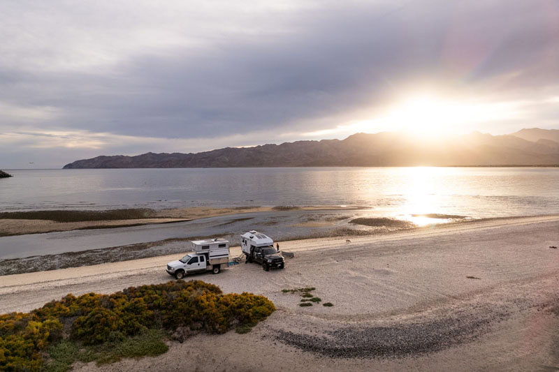Four Wheel Camper setups boondocking off-grid by serene ocean at sunrise over mountain silhouette.