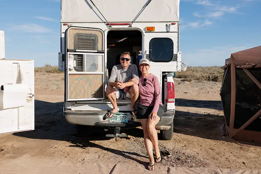 Couple sitting at open door of Four Wheel Camper boondocking off-grid, overlanding setup.