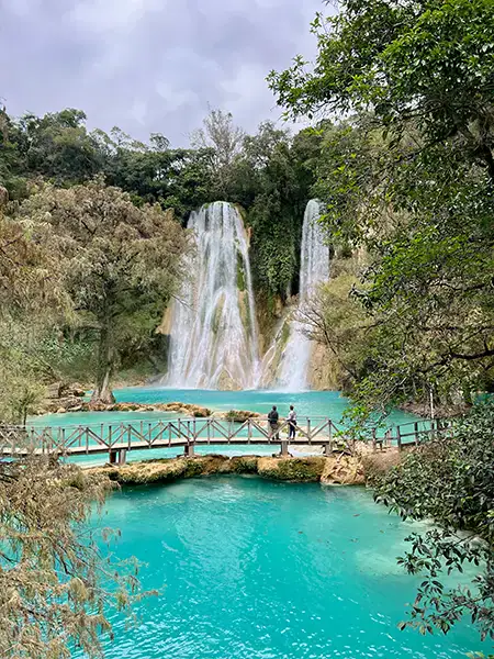Turquoise waterfall cascades over rock ledges, wooden bridge connects islands, two people observe nature.